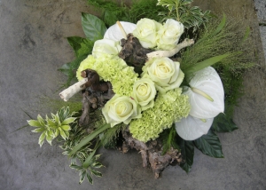 Een sober rouwbloemstukje met witte rozen, Anthurium en anjers je op basis van druivenhou, witte Avalanche rozen, Anthurium en groene anjers afgewerkt met klimop blad en grassen